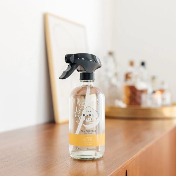 A clear spray bottle with a black nozzle is prominently displayed on a wooden surface. The label on the bottle reads "THE BARE HOME" and indicates it contains an all-purpose cleaner with a lemon tea tree scent. The bottle holds 500 ml and features a simple, elegant design. In the background, there are blurred glass containers and a framed artwork, adding a cozy ambiance to the scene.