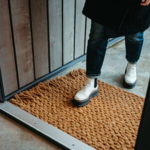 A person is stepping onto a textured, brown doormat made of thick, woven fibers. They are wearing white ankle boots with a chunky black sole. The background features wooden paneling and a concrete floor, suggesting an indoor entrance area. The jeans are dark and slightly rolled at the cuffs.