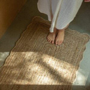 A textured, woven rug with a scalloped edge lies on a smooth, light-colored floor. Bare feet are positioned on the rug, which is made of natural fibers, suggesting a cozy and inviting atmosphere. Soft, natural light filters in, casting gentle shadows on the floor and rug, enhancing the serene ambiance of the space. The walls are a warm wood tone, contributing to the overall warmth of the scene.