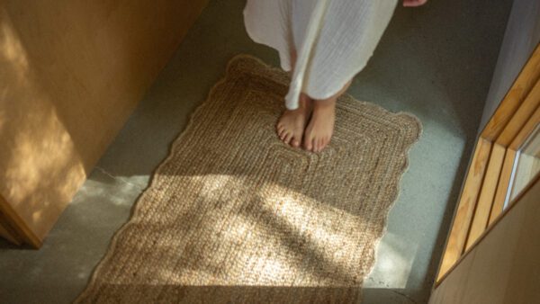 A textured, woven rug with a scalloped edge lies on a smooth, light-colored floor. Bare feet are positioned on the rug, which is made of natural fibers, suggesting a cozy and inviting atmosphere. Soft, natural light filters in, casting gentle shadows on the floor and rug, enhancing the serene ambiance of the space. The walls are a warm wood tone, contributing to the overall warmth of the scene.