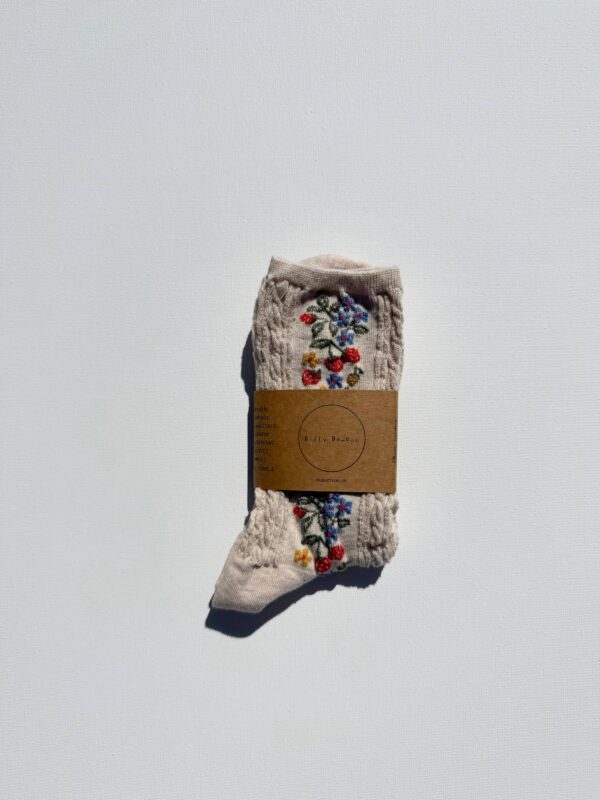 A single pair of light beige socks is displayed against a white background. The socks feature a textured knit pattern and are adorned with colorful floral embroidery in red, blue, and yellow. They are secured with a brown paper tag that includes the brand name "Billy Bamboo" and additional product information.