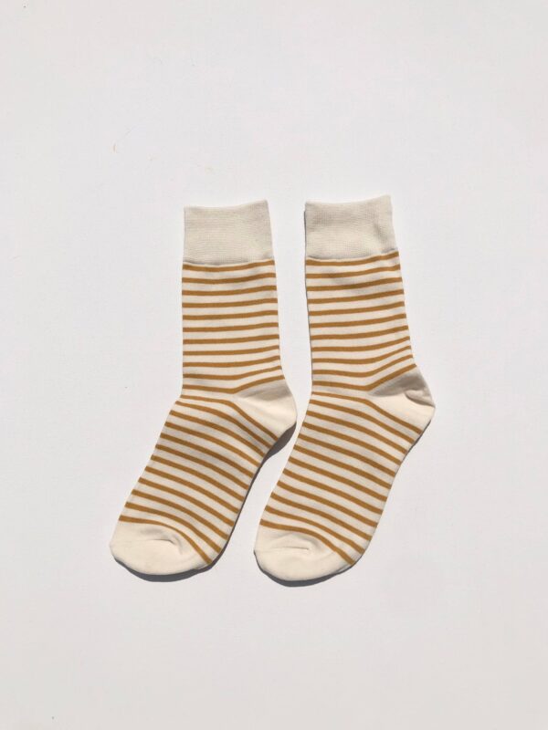 A pair of socks is displayed, featuring a cream base color with alternating thin and thick stripes in a warm, golden brown. The socks have a solid cream cuff at the top and a rounded toe design. They are positioned side by side on a plain white background.