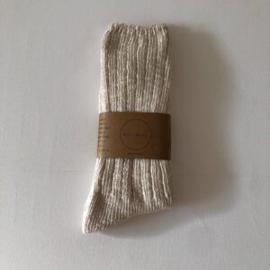 A pair of light beige, ribbed socks is neatly folded and secured with a brown paper band. The band features text that includes the brand name and care instructions. The socks have a soft, textured appearance, and the background is a plain white surface.