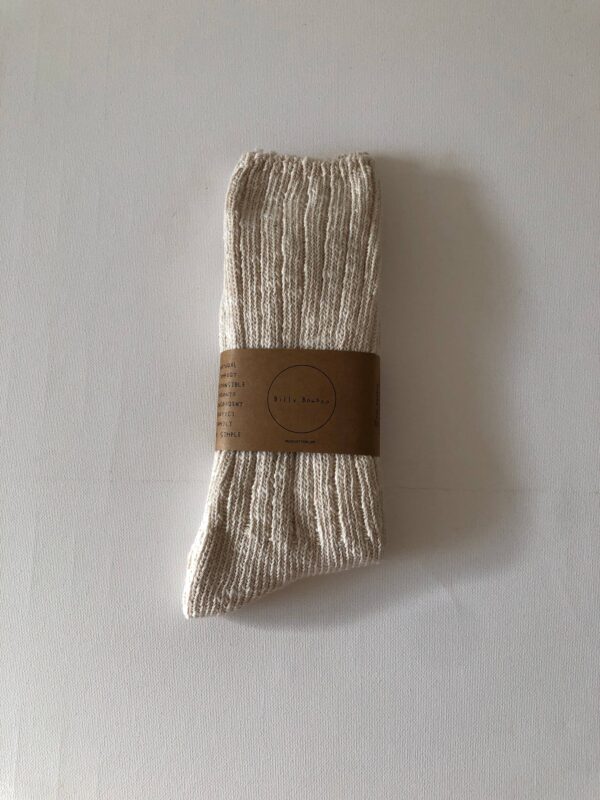 A pair of light beige, ribbed socks is neatly folded and secured with a brown paper band. The band features text that includes the brand name and care instructions. The socks have a soft, textured appearance, and the background is a plain white surface.