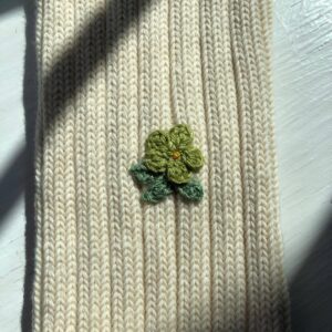 A knitted fabric in a light cream color features a small, embroidered flower design. The flower has four petals in a vibrant green hue, with a yellow center, and is accompanied by two green leaves. The texture of the knit is visible, showcasing a ribbed pattern. Soft shadows from nearby objects create a gentle contrast on the surface.