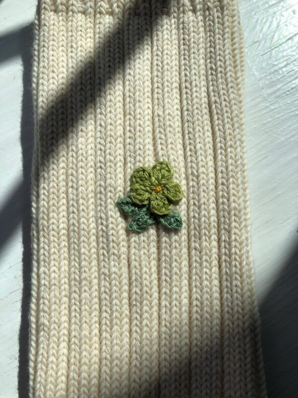 A knitted fabric in a light cream color features a small, embroidered flower design. The flower has four petals in a vibrant green hue, with a yellow center, and is accompanied by two green leaves. The texture of the knit is visible, showcasing a ribbed pattern. Soft shadows from nearby objects create a gentle contrast on the surface.