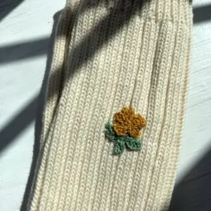 A close-up of a light beige knitted fabric, featuring a small embroidered flower design. The flower has a golden-yellow center with green leaves beneath it. The texture of the knit is visible, showcasing a ribbed pattern. The fabric is positioned on a white surface, with soft shadows cast across it.