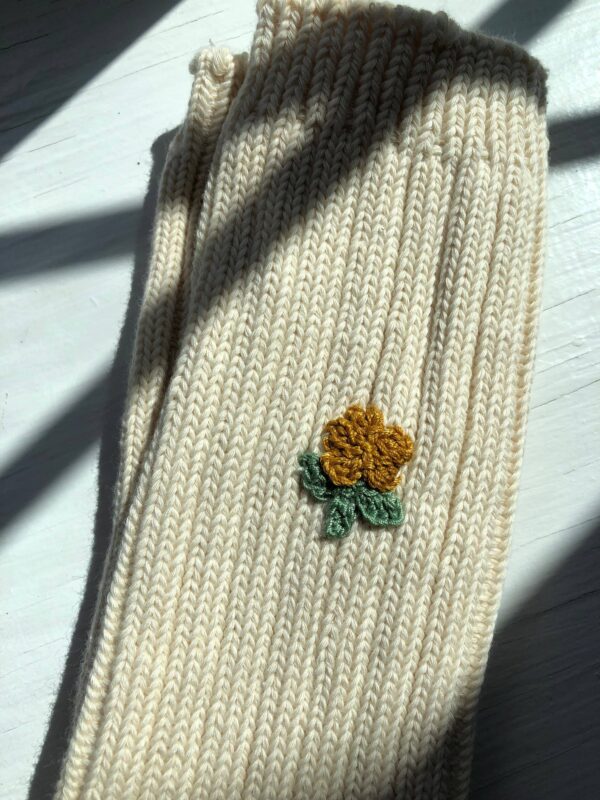A close-up of a cream-colored knitted fabric, featuring a small embroidered design of a yellow flower with green leaves. The texture of the knit is visible, and the fabric is laid flat against a light surface, with shadows creating a contrast.