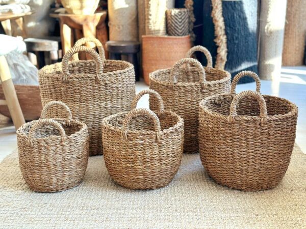 A collection of five woven baskets made from natural fibers is displayed. The baskets vary in size, with two smaller ones on the left, a medium-sized basket in the center, and two larger baskets on the right. Each basket features a rounded shape and two sturdy handles for easy carrying. The texture of the woven material is visible, showcasing a natural, earthy tone. The background includes additional woven items, adding to the rustic aesthetic. The baskets are placed on a light-colored rug, enhancing their natural look.