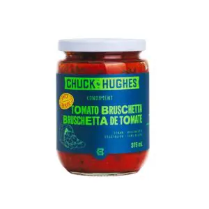 A glass jar of tomato bruschetta condiment is displayed. The jar features a white lid and a vibrant label with a blue background. The label prominently displays the brand name "Chuck Hughes" in white and green text, along with the product name "Tomato Bruschetta" in bold green letters. Additional details indicate that the product is vegan and gluten-free. The jar contains a rich, red mixture, suggesting a flavorful tomato-based condiment.