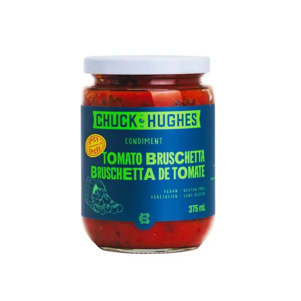 A glass jar of tomato bruschetta condiment is displayed. The jar features a white lid and a vibrant label with a blue background. The label prominently displays the brand name "Chuck Hughes" in white and green text, along with the product name "Tomato Bruschetta" in bold green letters. Additional details indicate that the product is vegan and gluten-free. The jar contains a rich, red mixture, suggesting a flavorful tomato-based condiment.