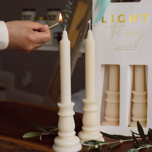 A hand is holding a lit matchstick, poised to light one of two tall, cream-colored candles on a wooden surface. The candles are placed in elegant holders with a classic design. In the background, a white packaging labeled "LIGHT the way" is visible, containing additional candles. Olive branches are scattered around the base, adding a natural touch to the scene.